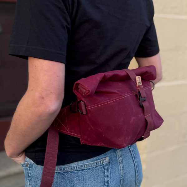Person wearing a black shirt and blue jeans with a red bag over their shoulder, standing on a sidewalk.