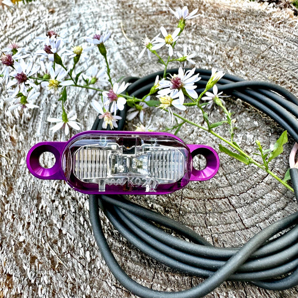 A purple and clear LED light bar with attached black cable, photographed on a weathered wood surface with small white flowers placed around it.