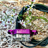 A purple SON brand bicycle headlight with a clear lens, resting on a coiled black cable over a textured wood surface with small white flowers.