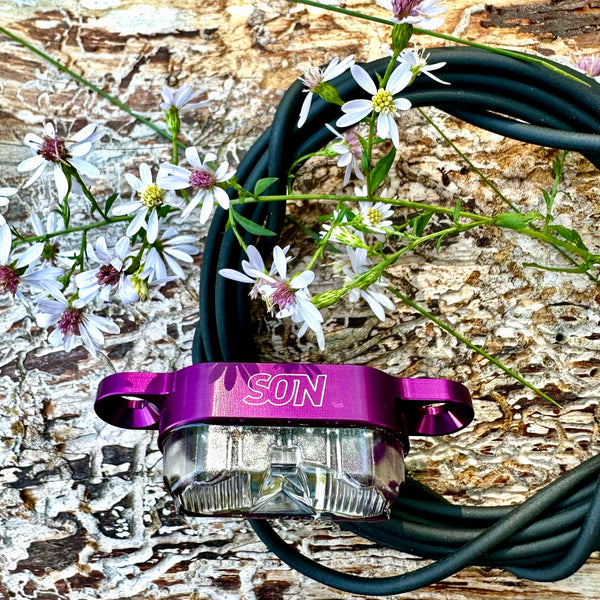 A purple SON brand bicycle headlight with a clear lens, resting on a coiled black cable over a textured wood surface with small white flowers.