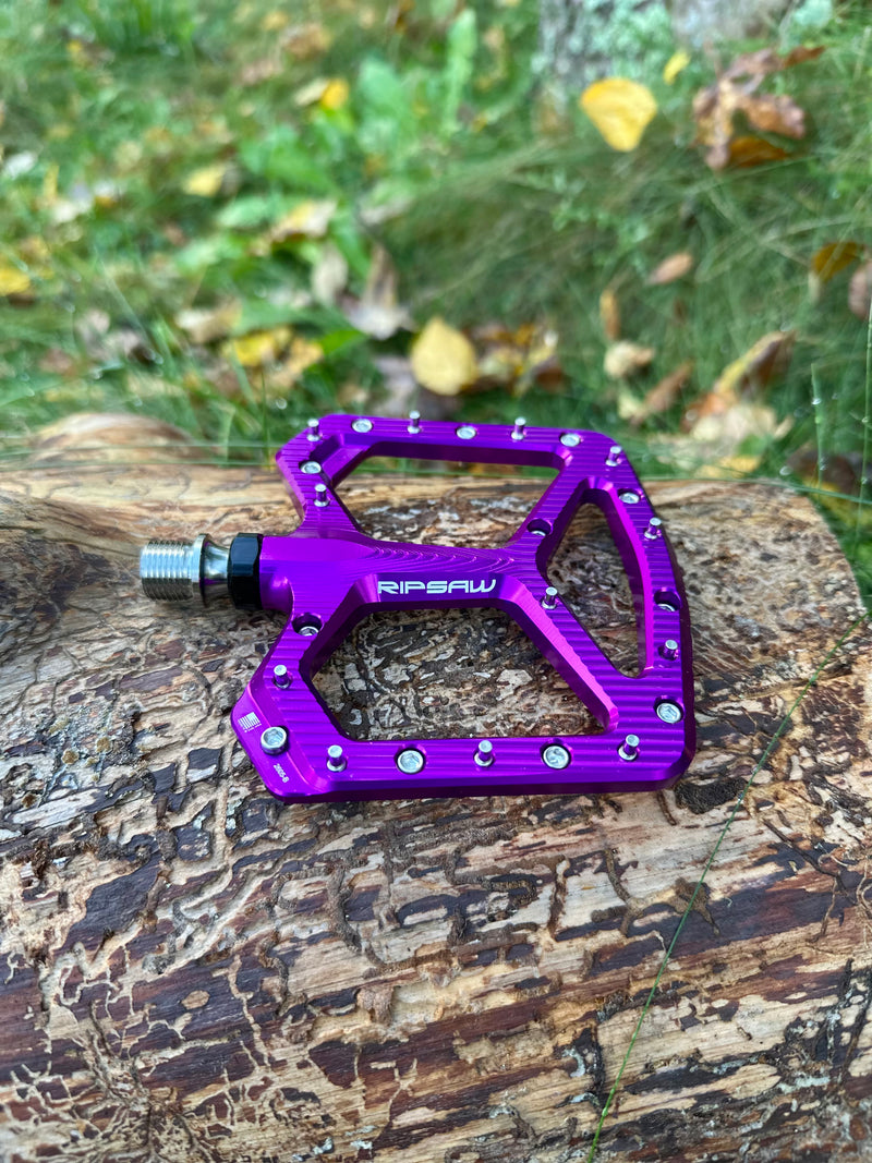 A bright purple anodized bicycle pedal with silver rivets and the model  name 'RIPSAW' printed on the side, resting on a textured log with a blurred outdoor grassy background.